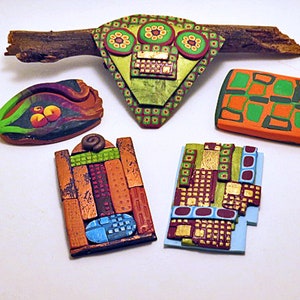 Puede incluir: Cinco colgantes de arcilla polimérica coloridos con diseños geométricos y florales. Los colgantes están dispuestos en una rama de madera y una superficie blanca.