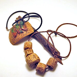 Pu&ograve; includere: Una collana caratterizzata da un pezzo marrone curvo e testurizzato con tre accenti dipinti in verde e rosso, abbinato a un cordino marrone infilato con quattro perline di legno cilindriche e un elemento simile a un rametto scuro.