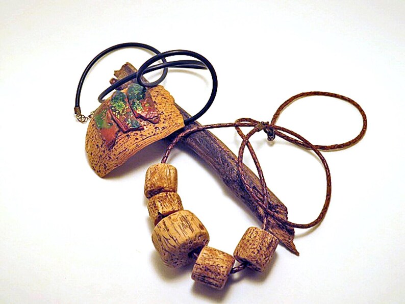 Pu&ograve; includere: Una collana caratterizzata da un pezzo marrone curvo e testurizzato con tre accenti dipinti in verde e rosso, abbinato a un cordino marrone infilato con quattro perline di legno cilindriche e un elemento simile a un rametto scuro.