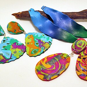 May include: A set of three pairs of colourful, abstract, dangle earrings. The earrings are made of polymer clay and feature a variety of colours, including blue, green, orange, yellow, pink, and purple. The earrings are all different shapes and sizes, and they are all very eye-catching.