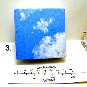 May include: A small blue canvas with white paint depicting clouds. The canvas is 3 inches wide and 2 inches tall.