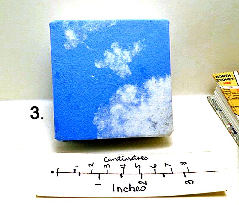 May include: A small blue canvas with white paint depicting clouds. The canvas is 3 inches wide and 2 inches tall.