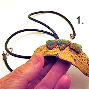 Pu&ograve; includere: Una collana con un ciondolo curvo e testurizzato color marrone chiaro con una superficie porosa. Tre piccoli pezzi triangolari screziati di verde e marrone sono fissati sulla parte superiore. Il ciondolo pende da un sottile cordino nero con chiusura.