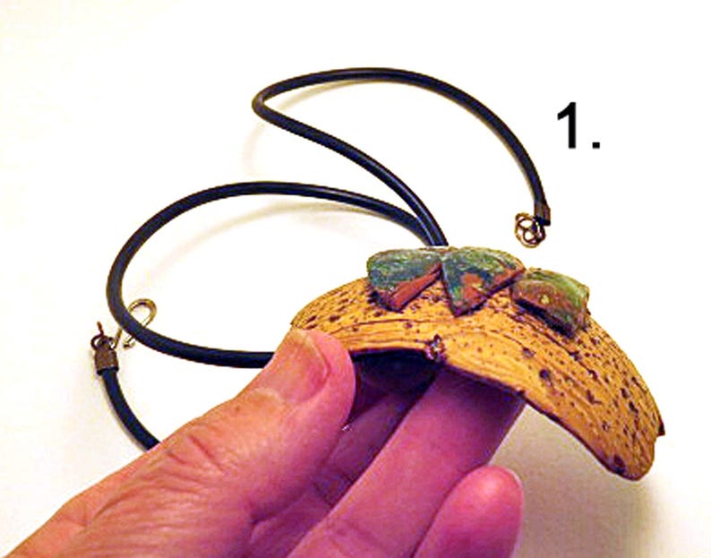 Pu&ograve; includere: Una collana con un ciondolo curvo e testurizzato color marrone chiaro con una superficie porosa. Tre piccoli pezzi triangolari screziati di verde e marrone sono fissati sulla parte superiore. Il ciondolo pende da un sottile cordino nero con chiusura.