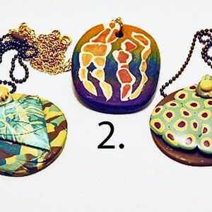 May include: Three circular pendants on ball chains. Pendant 1 is brown with a green and yellow spotted overlay. Pendant 2 is purple with a rainbow gradient and white-outlined orange shapes. Pendant 3 features a green, blue, and brown camouflage pattern.