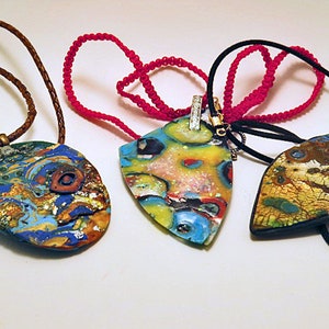 May include: Three colourful necklaces with unique pendant designs. The pendants are made of glass and feature abstract patterns in shades of blue, green, yellow, and brown. The necklaces are strung on different types of cord, including leather, string, and a black elastic cord.