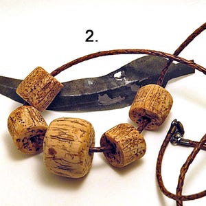 Pu&ograve; includere: Una collana con cinque perline cilindriche di colore marrone chiaro e dalla superficie ruvida, infilate su un cordino intrecciato marrone scuro. Il numero 2 &egrave; stampato sopra la collana, posta davanti a un oggetto metallico scuro e curvo.