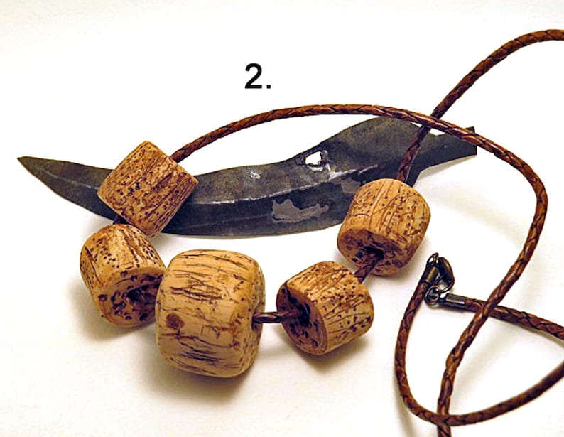 Pu&ograve; includere: Una collana con cinque perline cilindriche di colore marrone chiaro e dalla superficie ruvida, infilate su un cordino intrecciato marrone scuro. Il numero 2 &egrave; stampato sopra la collana, posta davanti a un oggetto metallico scuro e curvo.