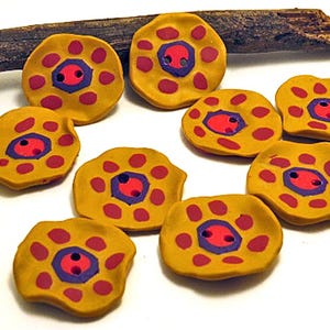Peut inclure: Un ensemble de neuf boutons ronds jaune moutarde aux bords irréguliers et ondulés. Chaque bouton présente un hexagone violet central avec deux trous, entouré d'un cercle de petits points circulaires rose foncé disposés en motif floral.