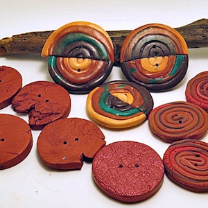 Könnte beinhalten: Sortiment handgefertigter Tonknöpfe in verschiedenen Farben und Mustern, darunter Braun, Rot, Grün und Gelb. Einige Knöpfe haben ein Spiraldesign, während andere ein rustikaleres, strukturiertes Aussehen haben.