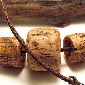 Pu&ograve; includere: Una collana con diverse perline color marrone chiaro, cilindriche e cubiche, con una superficie porosa e strutturata. Le perline sono infilate su un sottile cordino di cuoio intrecciato marrone scuro che passa sopra la perla centrale e include una chiusura in metallo.