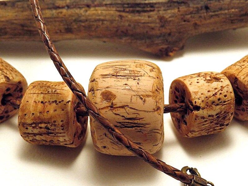 Pu&ograve; includere: Una collana con diverse perline color marrone chiaro, cilindriche e cubiche, con una superficie porosa e strutturata. Le perline sono infilate su un sottile cordino di cuoio intrecciato marrone scuro che passa sopra la perla centrale e include una chiusura in metallo.