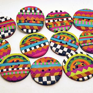 May include: A collection of ten round, two-hole buttons featuring vibrant, multi-colored geometric patterns. Each button displays a unique arrangement of stripes, zigzags, and checkerboard designs in bright shades of pink, orange, blue, and green.