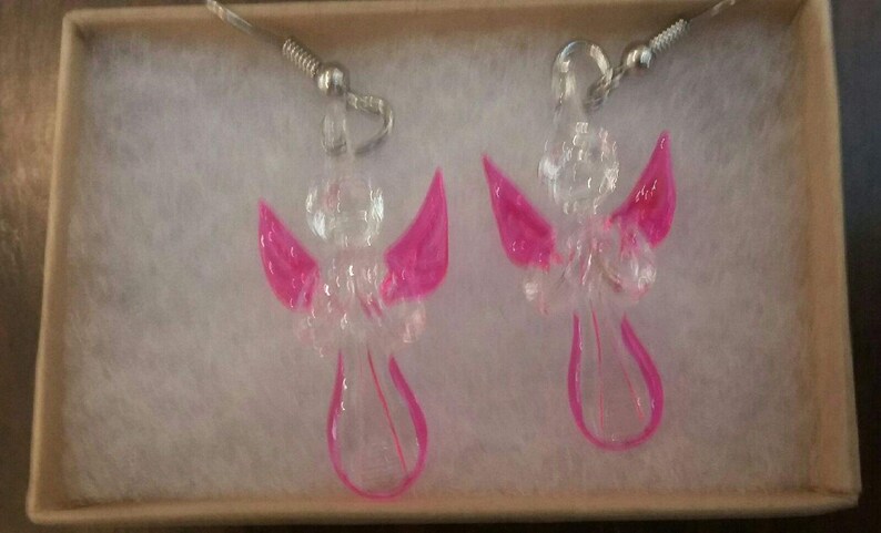 Puede incluir: Un par de pendientes de &aacute;ngel de vidrio transparente con alas rosas. Los pendientes cuelgan de ganchos plateados.