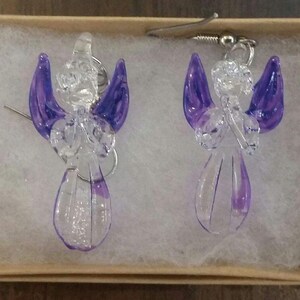 Puede incluir: Un par de pendientes de &aacute;ngel de vidrio con alas moradas. Los pendientes est&aacute;n en una peque&ntilde;a caja con papel de seda blanco.