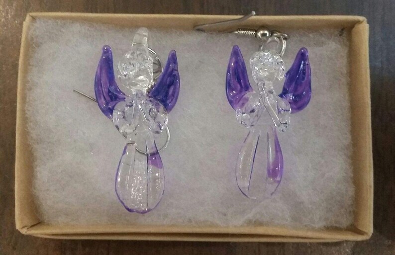 Puede incluir: Un par de pendientes de &aacute;ngel de vidrio con alas moradas. Los pendientes est&aacute;n en una peque&ntilde;a caja con papel de seda blanco.