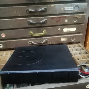 K&ouml;nnte beinhalten: Ein schwarzes, ledergebundenes Buch mit einem Stern- und Halbmond-Design auf dem Einband. Das Buch liegt auf einem Holzschubladenkasten mit Metallgriffen und Etiketten.