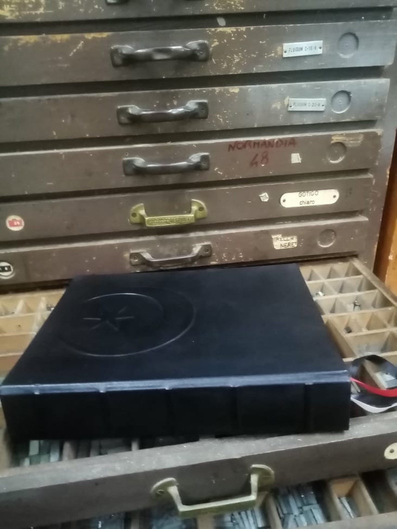 K&ouml;nnte beinhalten: Ein schwarzes, ledergebundenes Buch mit einem Stern- und Halbmond-Design auf dem Einband. Das Buch liegt auf einem Holzschubladenkasten mit Metallgriffen und Etiketten.