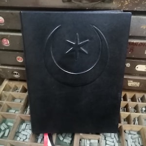 K&ouml;nnte beinhalten: Schwarzes, ledergebundenes Tagebuch mit einem gepr&auml;gten silbernen Halbmond- und Sternmuster auf dem vorderen Umschlag. Das Tagebuch ist geschlossen und liegt auf einer Holzoberfl&auml;che.