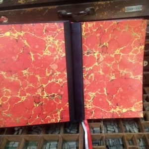 K&ouml;nnte beinhalten: Ein rotes und goldenes marmoriertes Buch mit schwarzem R&uuml;cken und einem roten, wei&szlig;en und schwarzen Lesezeichenband. Das Buch liegt auf einem Holztablett gef&uuml;llt mit Bleisatz.