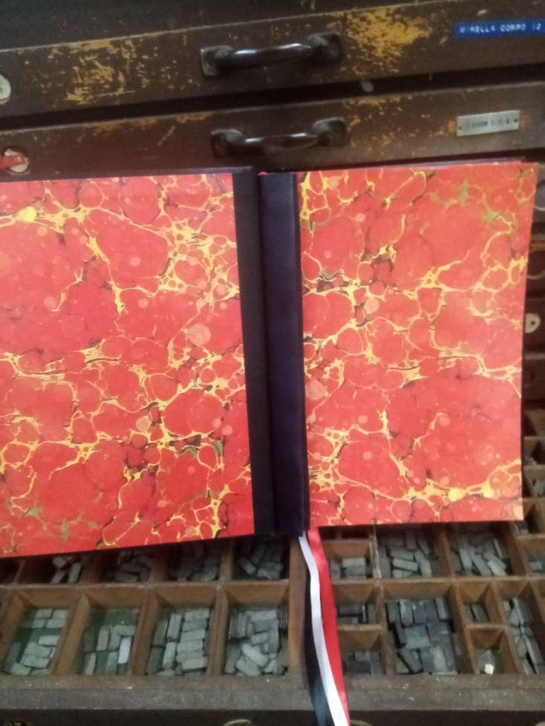 K&ouml;nnte beinhalten: Ein rotes und goldenes marmoriertes Buch mit schwarzem R&uuml;cken und einem roten, wei&szlig;en und schwarzen Lesezeichenband. Das Buch liegt auf einem Holztablett gef&uuml;llt mit Bleisatz.