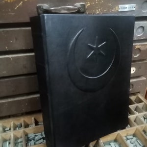 K&ouml;nnte beinhalten: Ein schwarzes, ledergebundenes Buch mit einem gepr&auml;gten Halbmond- und Sternmuster auf dem vorderen Einband. Das Buch liegt auf einer Holzoberfl&auml;che mit einer Schale aus Bleilettern.