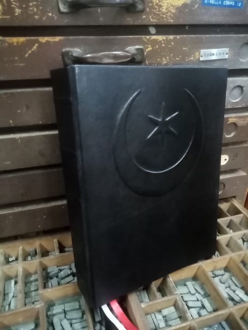 K&ouml;nnte beinhalten: Ein schwarzes, ledergebundenes Buch mit einem gepr&auml;gten Halbmond- und Sternmuster auf dem vorderen Einband. Das Buch liegt auf einer Holzoberfl&auml;che mit einer Schale aus Bleilettern.