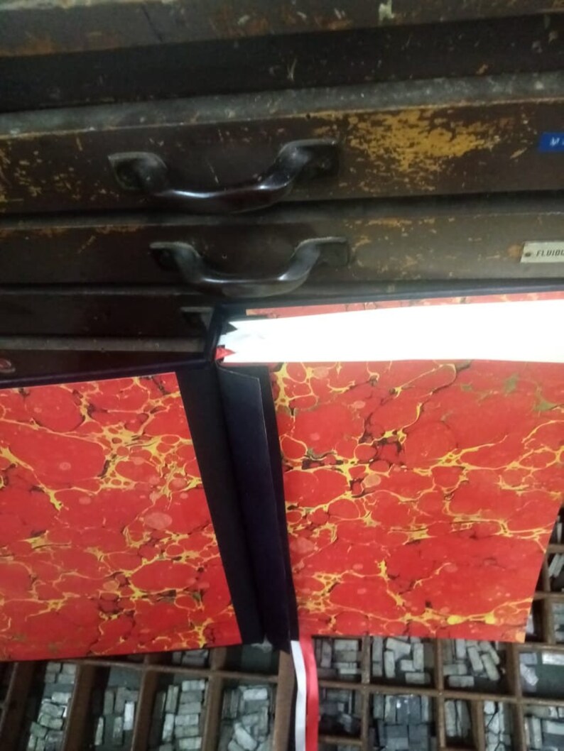 K&ouml;nnte beinhalten: Zwei rote und gold marmorierte Hardcover-B&uuml;cher mit schwarzen R&uuml;ckseiten und wei&szlig;en Band-Lesezeichen. Die B&uuml;cher liegen auf einer Oberfl&auml;che mit einem Raster aus Metalltypen.