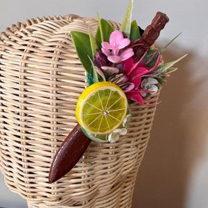 May include: A decorative hair accessory featuring a lemon slice, pink and red flowers, green leaves, and a brown tiki-style figure. The accessory is attached to a brown base and is set against a woven wicker background.