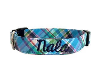 May include: A blue, green, and white plaid pet collar featuring the name Nala embroidered in dark blue script. The collar includes a black plastic side-release buckle, a black plastic adjuster, and a silver-toned metal D-ring for attachment.