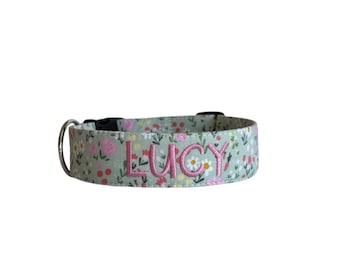 May include: A sage green pet collar featuring a repeating floral pattern with small white, pink, and yellow flowers and green foliage. The name LUCY is embroidered in pink capital letters on the front, with a black plastic buckle and metal D-ring.