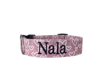 May include: A mauve pet collar featuring a repeating white heart pattern and the name Nala embroidered in dark thread. The collar includes a black plastic side-release buckle and a silver-toned metal D-ring for attaching a leash or identification tag.