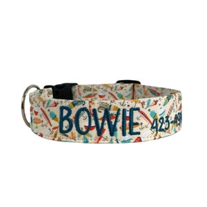 May include: A cream-colored dog collar with a pattern of fishing lures and tools. The collar has the name "BOWIE" and a phone number embroidered in navy blue. It features a black plastic buckle and a silver metal D-ring.