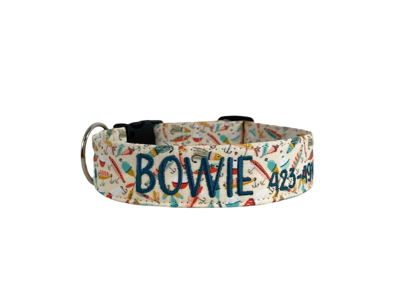 May include: A cream-colored dog collar with a pattern of fishing lures and tools. The collar has the name "BOWIE" and a phone number embroidered in navy blue. It features a black plastic buckle and a silver metal D-ring.