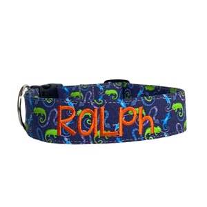 May include: A navy blue dog collar with a repeating pattern of green and blue chameleons. The name "RALPH" is embroidered in orange thread. The collar has a black plastic buckle and a silver metal D-ring.