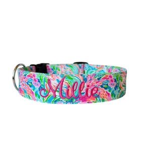 May include: A colorful dog collar with a floral pattern in shades of pink, blue, and green. The name "Millie" is embroidered in bright pink script across the front. The collar has a black plastic buckle and a silver metal D-ring.