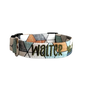 May include: A fabric pet collar featuring a geometric pattern in shades of orange, blue, and grey. The collar includes a silver metal D-ring, a black plastic buckle, and the name Walter embroidered in dark green thread on the front surface.