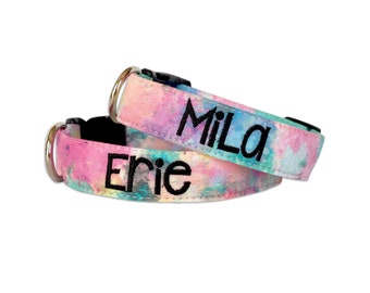 May include: Two pet collars featuring a watercolor-style pattern in shades of pink, blue, and yellow. Each collar has a black plastic buckle, a silver metal D-ring, and black embroidered text. One collar displays the name MiLa, and the other Erie.