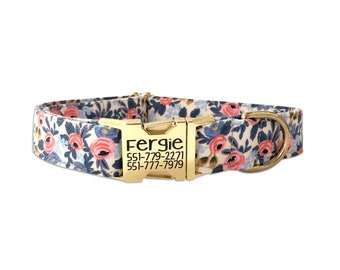 May include: A pet collar featuring a floral pattern with pink roses and blue leaves on a cream background. It includes a gold-toned metal buckle engraved with the name Fergie and two phone numbers: 551-779-2271 and 551-777-7979, plus a gold D-ring.