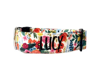 May include: A pet collar featuring a vibrant floral pattern with red, pink, yellow, and blue flowers on a cream background. The name LUCY is embroidered in bold black script across the center. It includes a black plastic buckle and a metal D-ring.