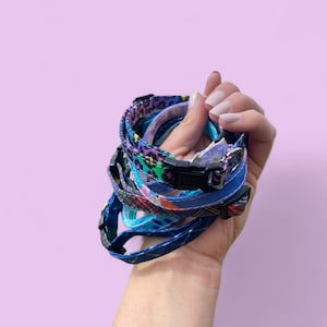 May include: A stack of colorful patterned cat collars with a black buckle. The collars are held in a hand.