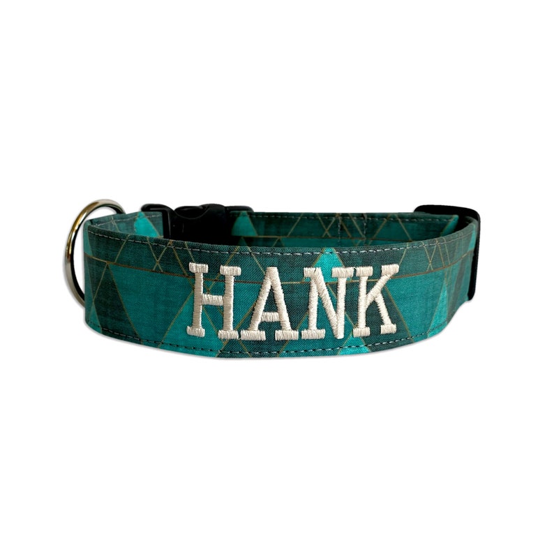 May include: A teal dog collar with a geometric pattern and the name "HANK" embroidered in white. The collar features a black buckle and a metal ring for leash attachment. The collar is designed for a pet.