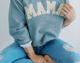 May include: A gray sweatshirt with the word "MAMA" in large, yellow letters with a floral pattern. The sweatshirt is worn by a person sitting on a wooden stool.