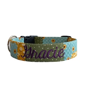 May include: A dog collar with a floral pattern in shades of yellow, blue, and green. The name "Gracie" is embroidered in purple thread on a green patch. The collar has a black plastic buckle and a silver metal ring.