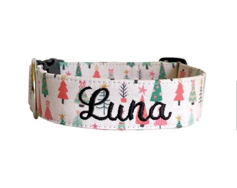 May include: A pet collar featuring a light-colored fabric with a repeating pattern of red, green, and white Christmas trees and stars. The name Luna is embroidered in black script lettering on the center of the collar, which includes a black buckle.