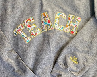 May include: Gray sweatshirt with the word "TEACH" in large, colorful letters with a school supply pattern. The letters are outlined in gold. The sleeve has the text "1st Grade" in gold embroidery.
