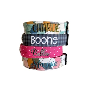 May include: A stack of four fabric dog collars with black plastic buckles. The top collar has a geometric pattern with the name CHARLIE embroidered in green. The second is navy plaid with BOONE in white. The third is pink with white dots and Bella in grey. The bottom has a geometric pattern with Maggie in blue.