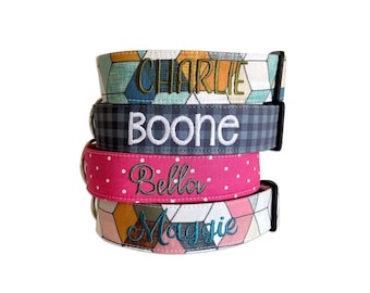 May include: A stack of four fabric dog collars with black plastic buckles. The top collar has a geometric pattern with the name CHARLIE embroidered in green. The second is navy plaid with BOONE in white. The third is pink with white dots and Bella in grey. The bottom has a geometric pattern with Maggie in blue.