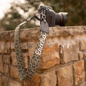 May include: A black camera with a patterned strap featuring a green and black leopard print. The strap is personalized with the name "Ericha" in white script. The camera rests on a stone wall.
