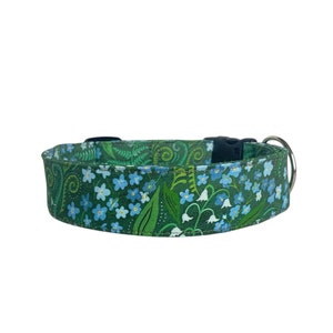 May include: A green fabric pet collar featuring a repeating pattern of small light blue flowers, white bell-shaped blossoms, and dark green fern leaves. The collar includes a black plastic side-release buckle and a silver metal D-ring attachment.